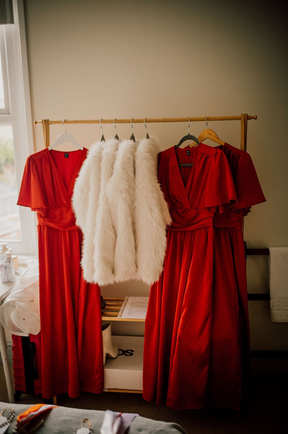 Red bridesmaid dresses and white fur shawls hanging on wooden racks in a dimly lit room.