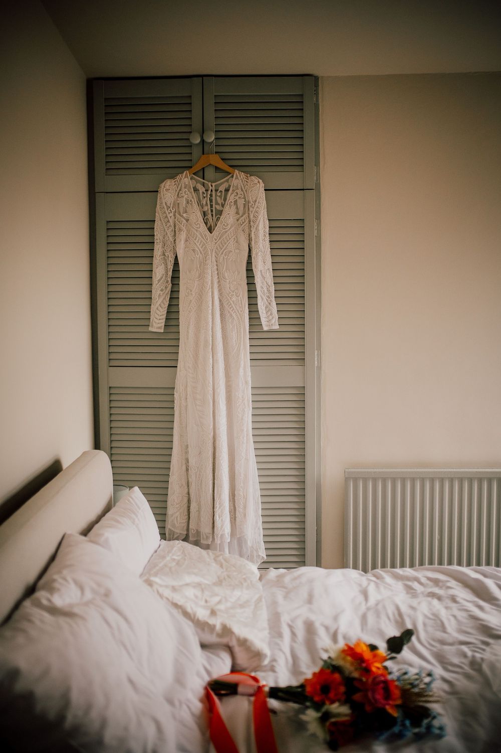 A vintage white lace wedding dress hangs elegantly against sage green shutters with red floral accents on a bed.