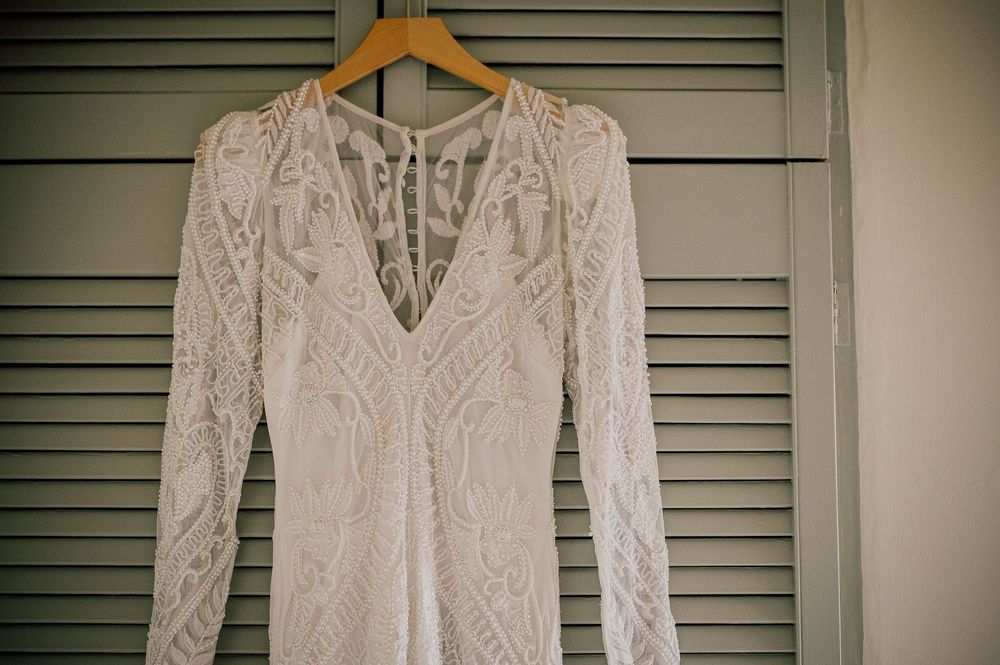 A white lace wedding dress with long sleeves hangs on a wooden hanger against green shutters.