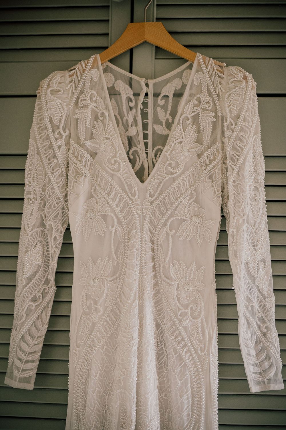 Elegant white lace wedding dress with long sleeves and intricate floral patterns hangs on wooden hanger against green shutters.