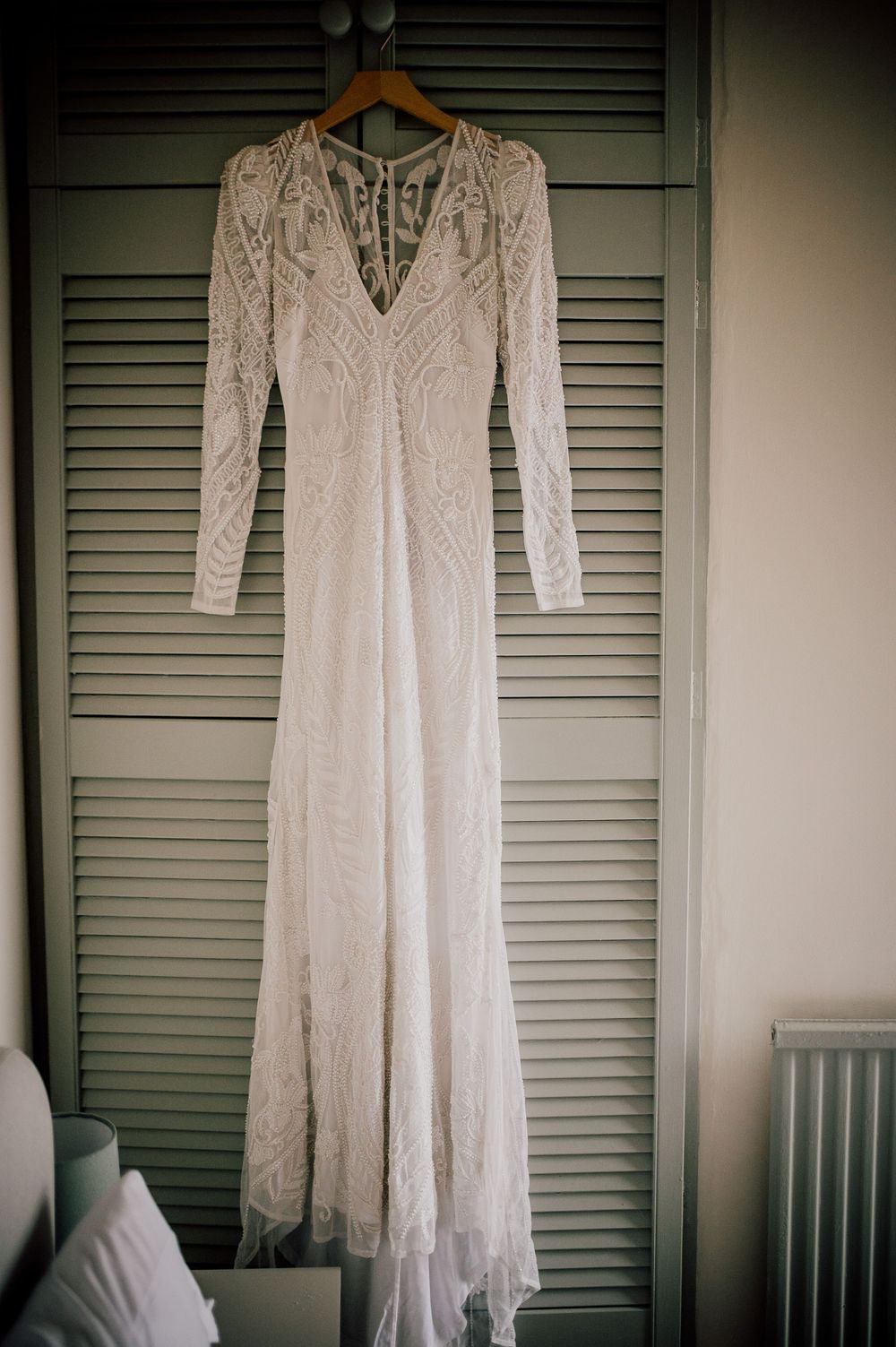 White lace wedding dress with long sleeves hanging on wooden hangers against gray shuttered wall.