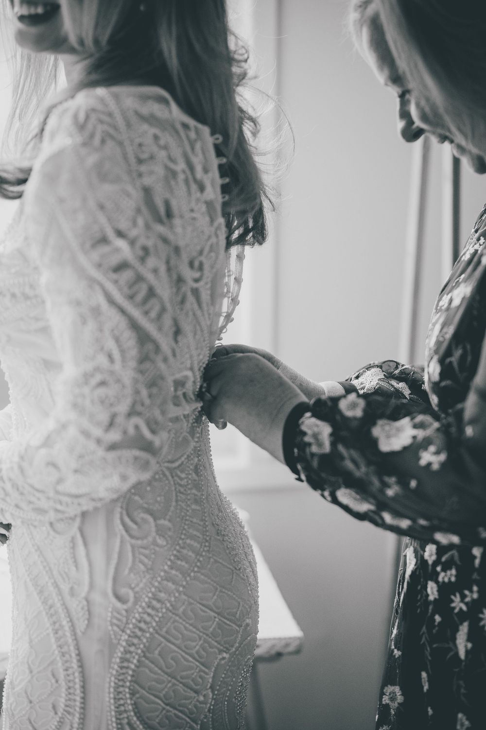 Black and white image of a bride in a detailed lace wedding dress being helped with final preparations.