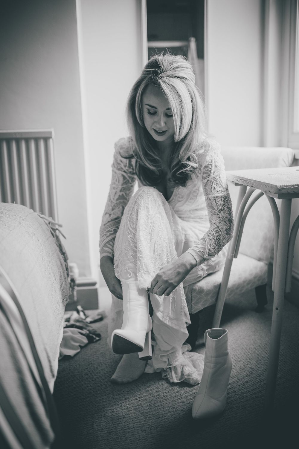 Black and white photo of a bride in a lace dress putting on white boots while sitting in a vintage style room.