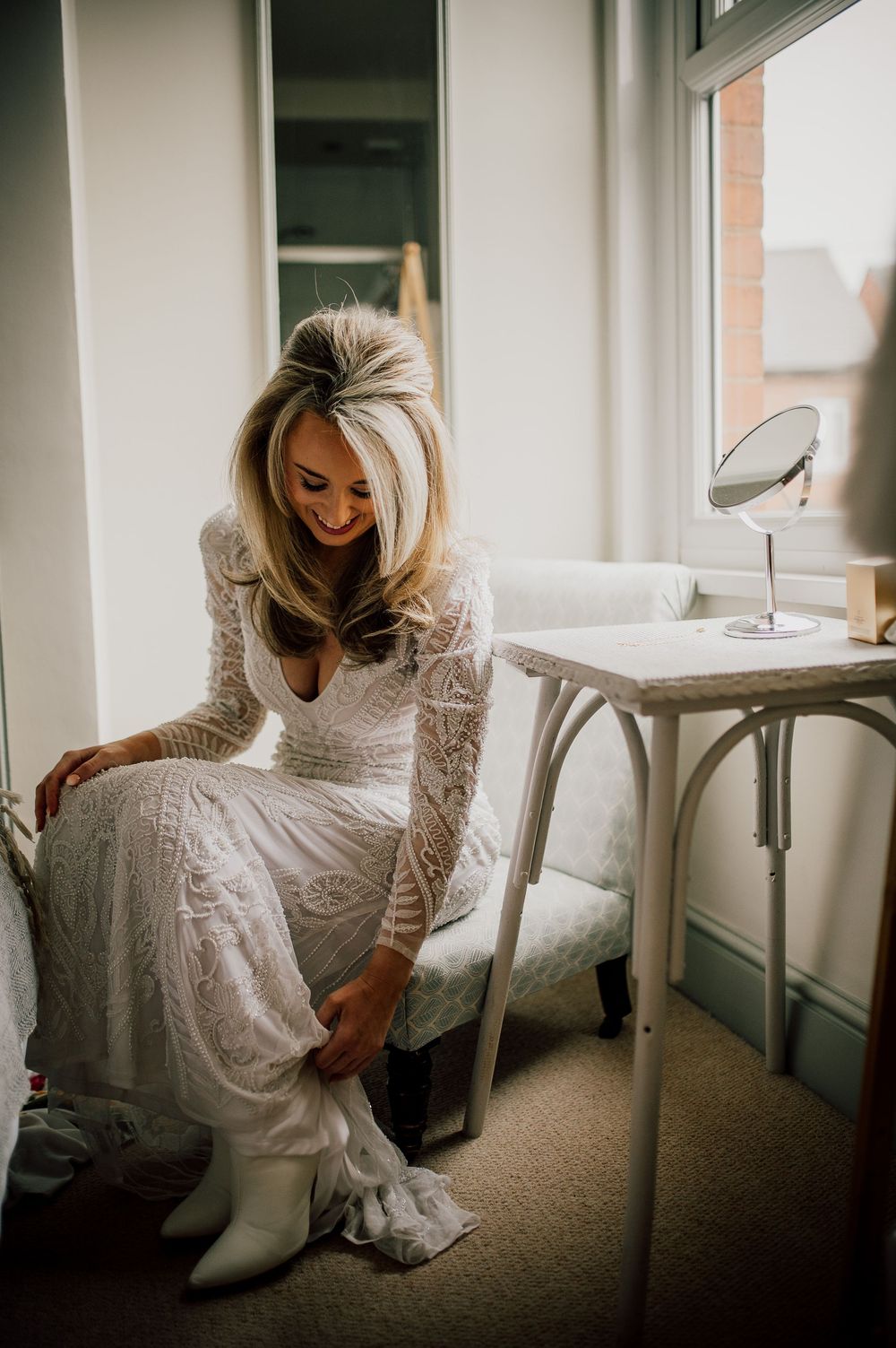 A bride in a white lace dress sits gracefully in a sunlit room with modern minimalist decor.