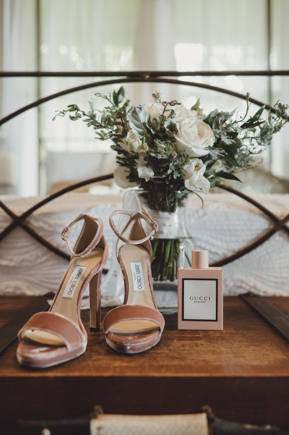 Elegant bridal shoes displayed with white roses and Gucci perfume on a wooden surface.