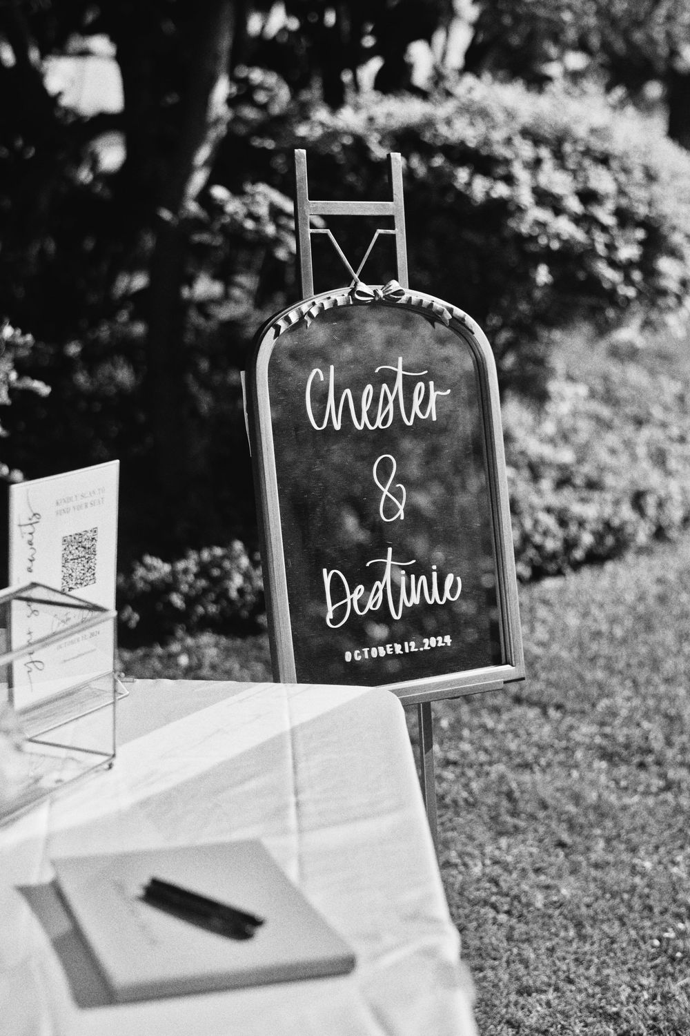 Black and white wedding signage displays elegant handwritten text on acrylic stands arranged on grass outdoors.
