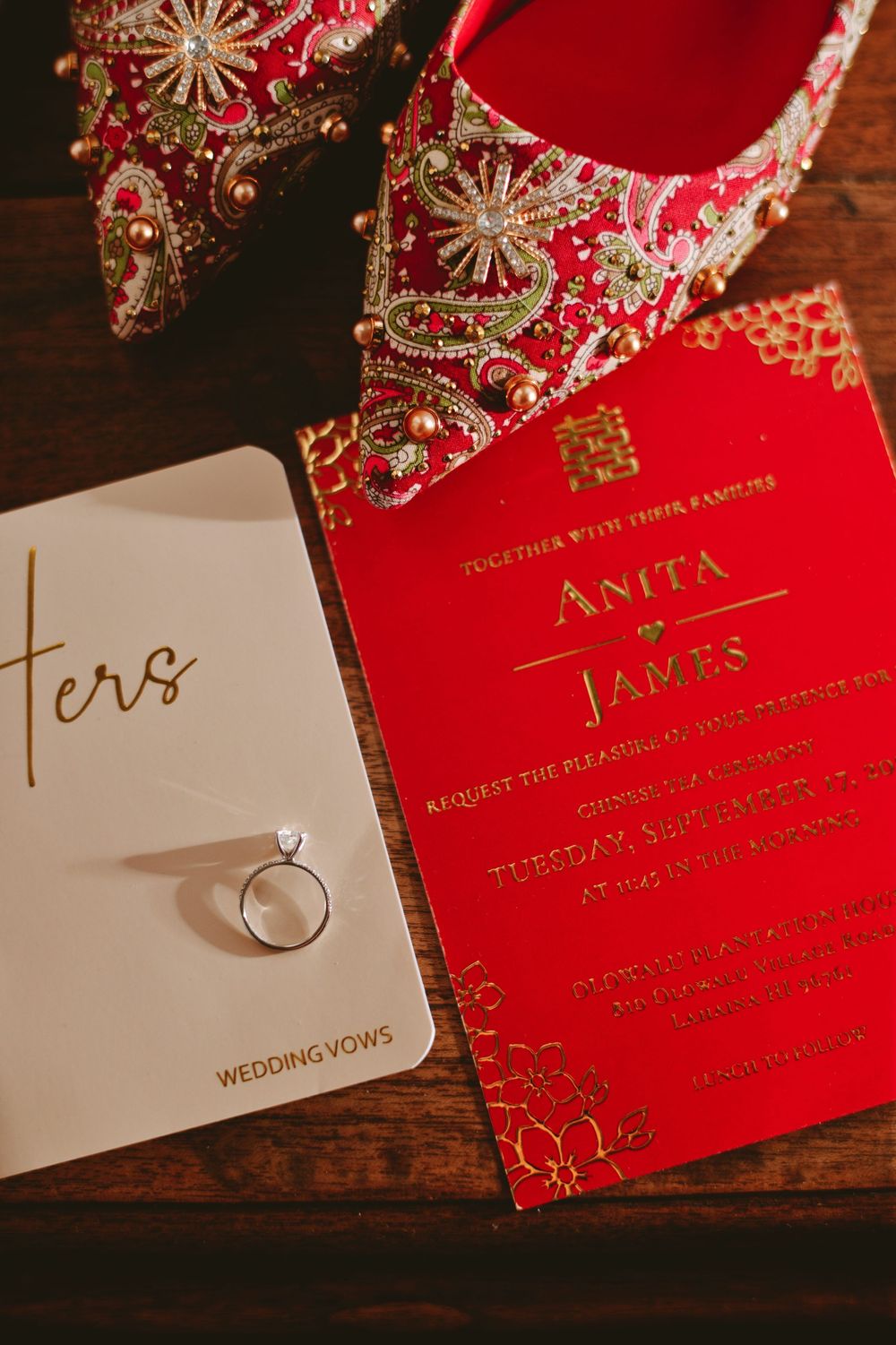 Elegant red and gold wedding invitation with ornate bridal shoes and engagement ring on wooden surface.