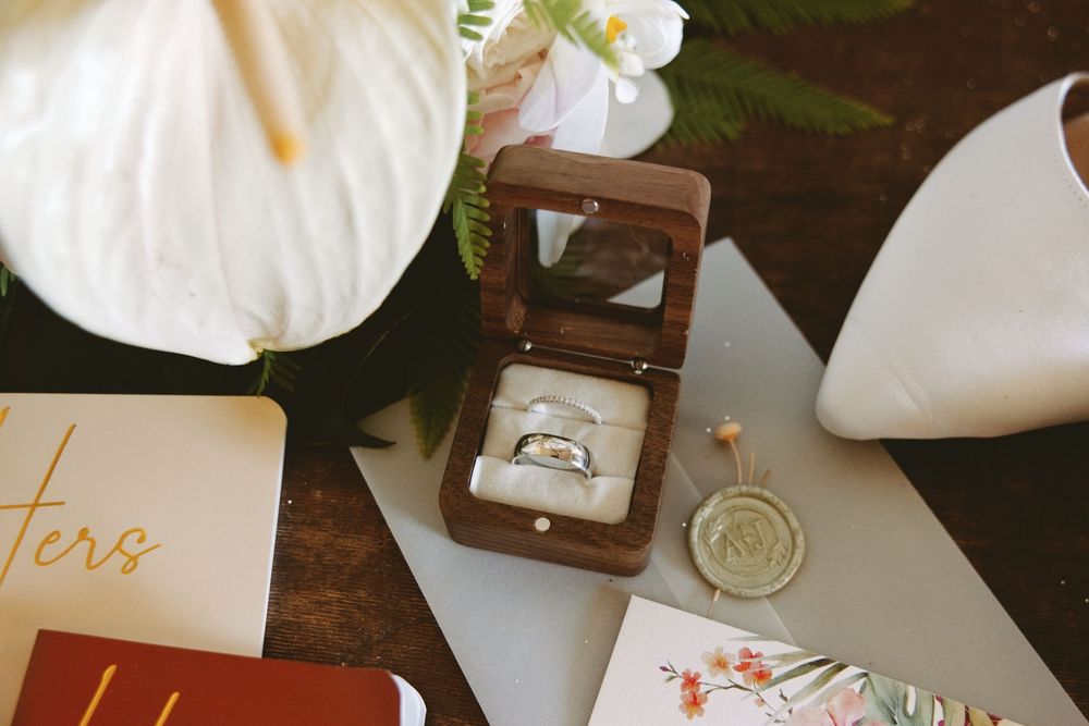 A collection of wedding details including white flowers, rings, invitations and decor arranged on a dark surface.