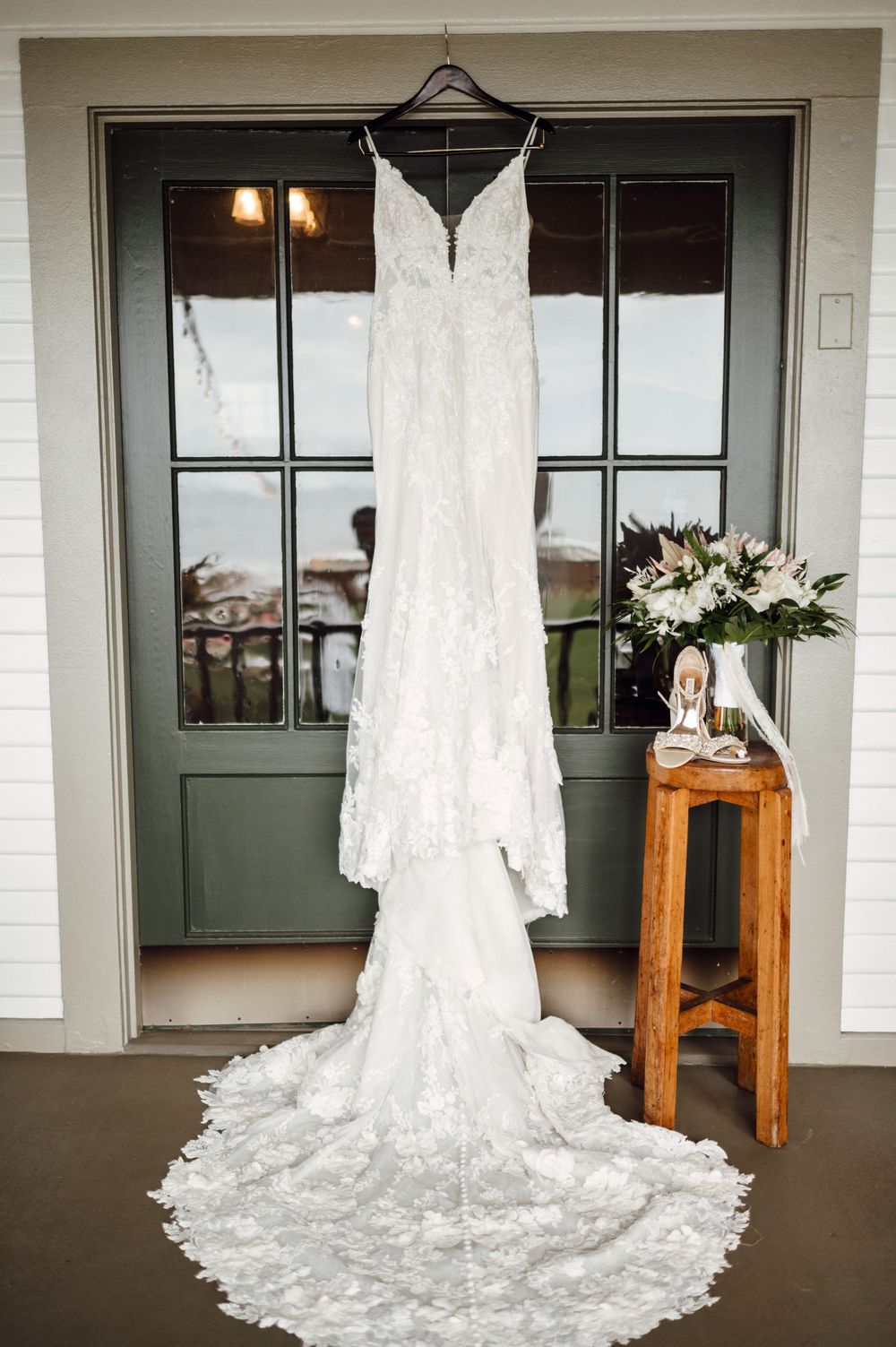 Elegant white lace wedding dress with long train hanging on display next to wooden side table with floral arrangement.
