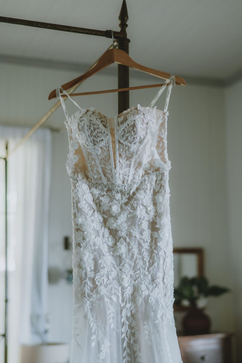 A delicate white lace wedding dress with intricate floral details hangs on a wooden hanger against a light wall.