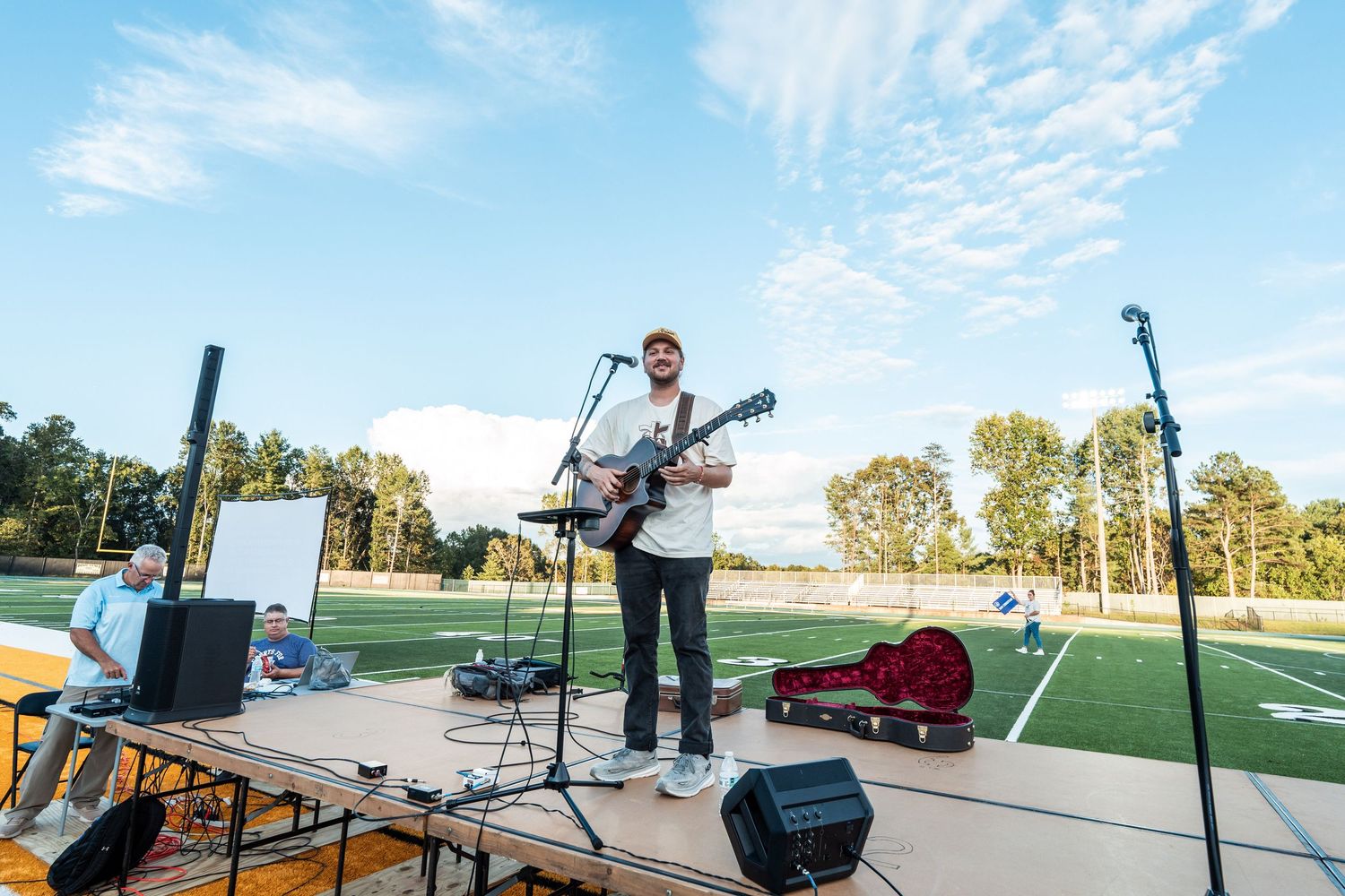 Musician performs with guitar on outdoor stage setup with sound equipment and instrument cases under blue sky.