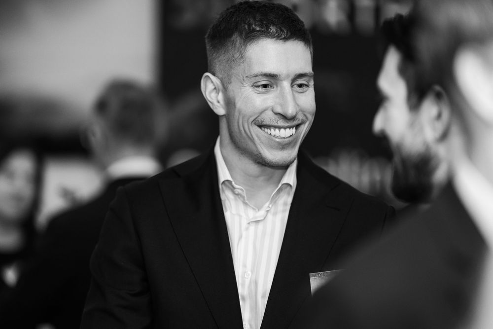 Black and white photo of a smiling person in a striped dress shirt and blazer at a social event.