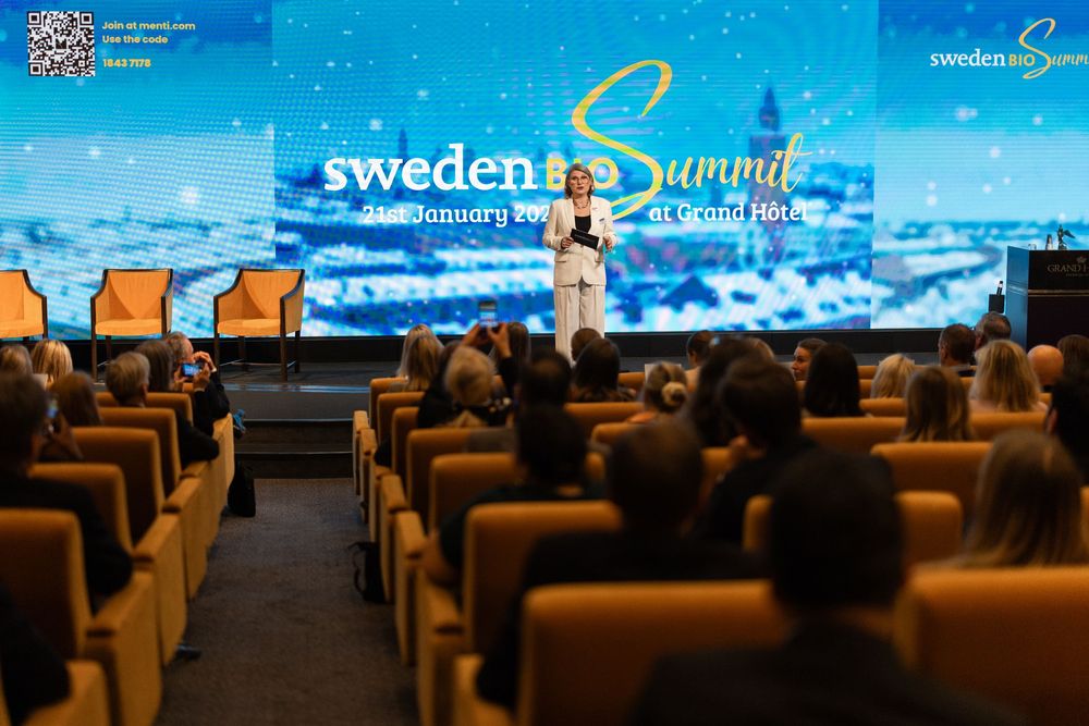 Conference attendees seated in rows facing a large blue-lit presentation screen during a business event.