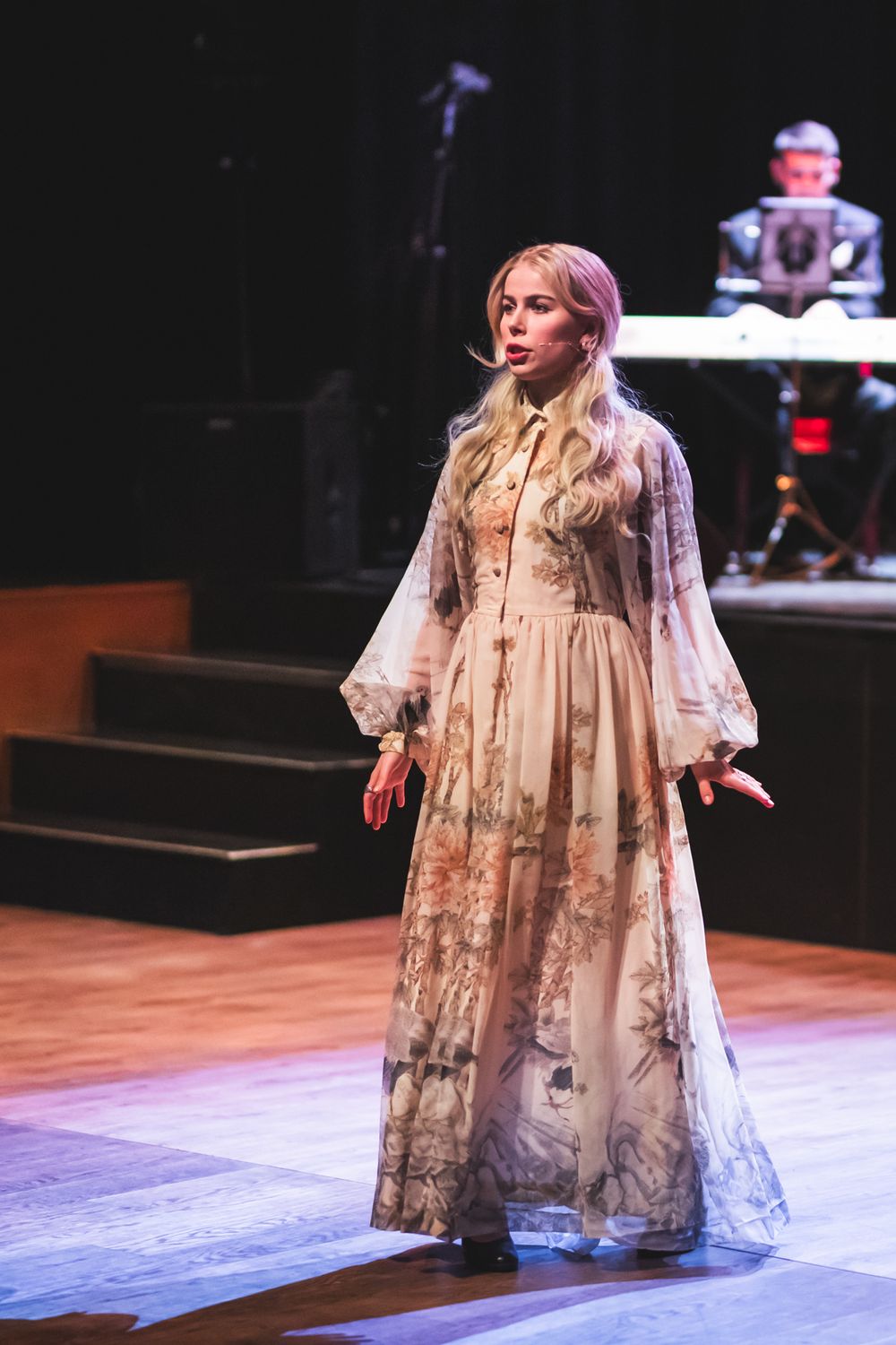 A performer in a flowing vintage-style cream dress with floral patterns sings on stage during a theatrical production.