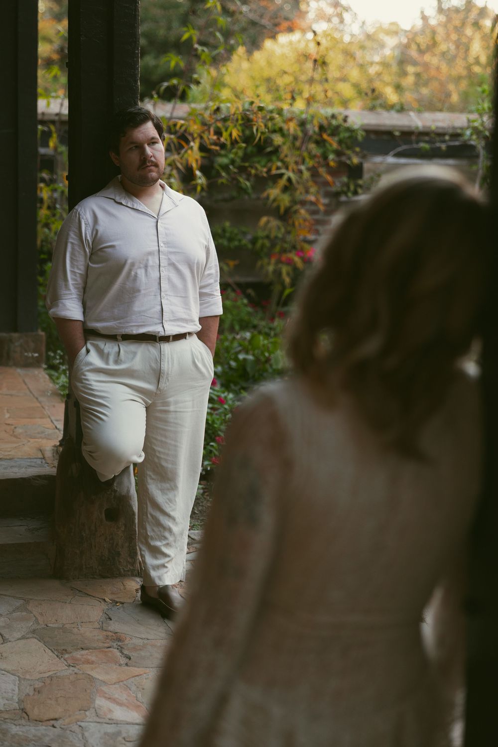 A couple in white outfits poses in a garden setting with stone pathways and greenery.