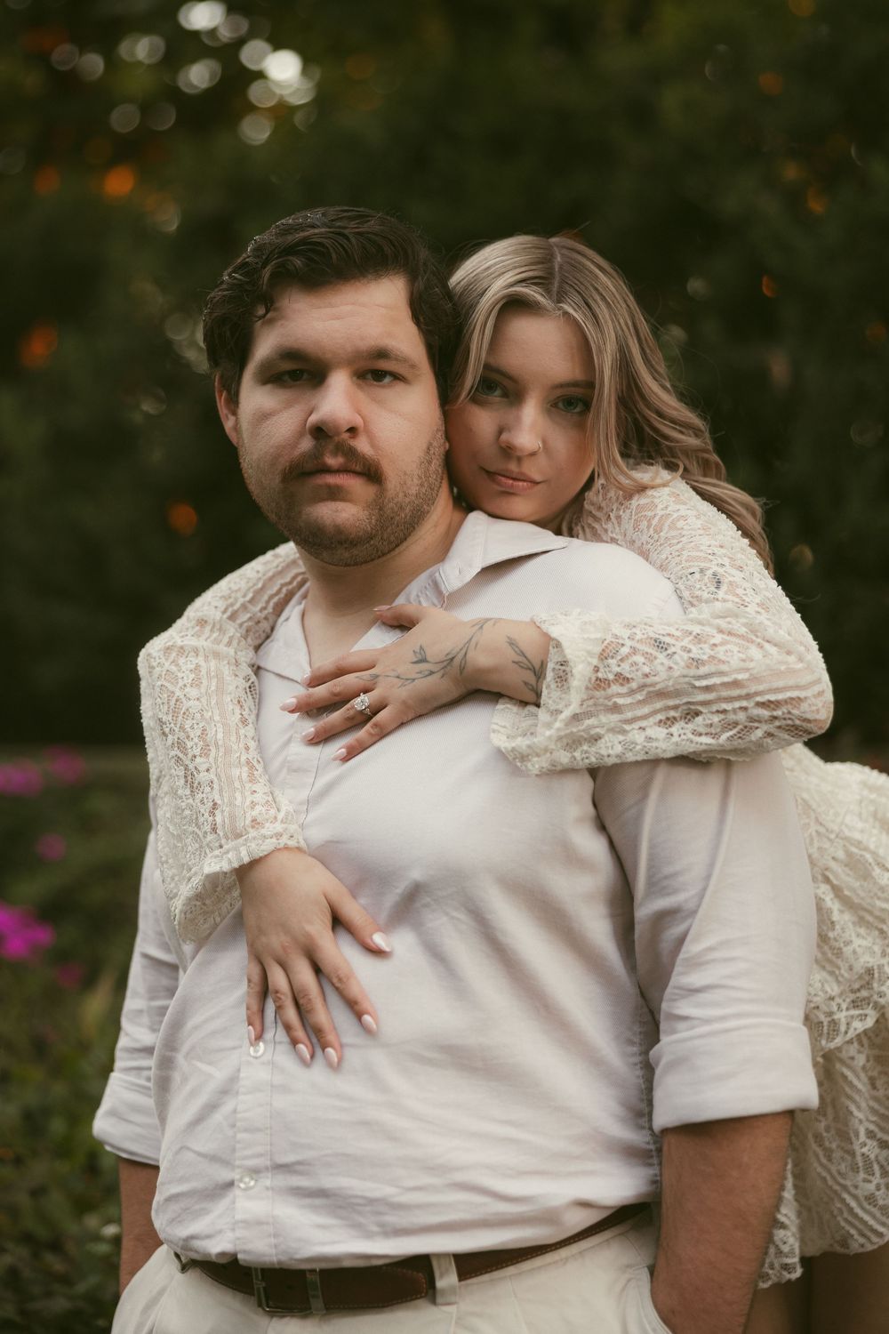 A couple in white embraces tenderly in a garden with blooming flowers.