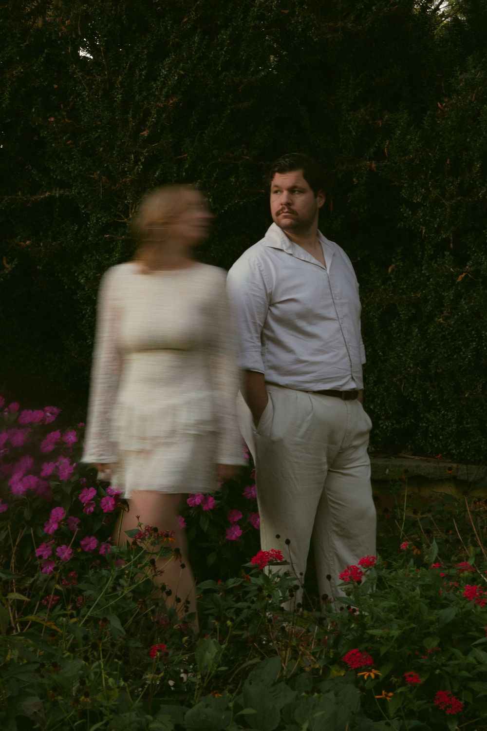 A couple in light clothing walks through a garden with vibrant pink and red flowers.