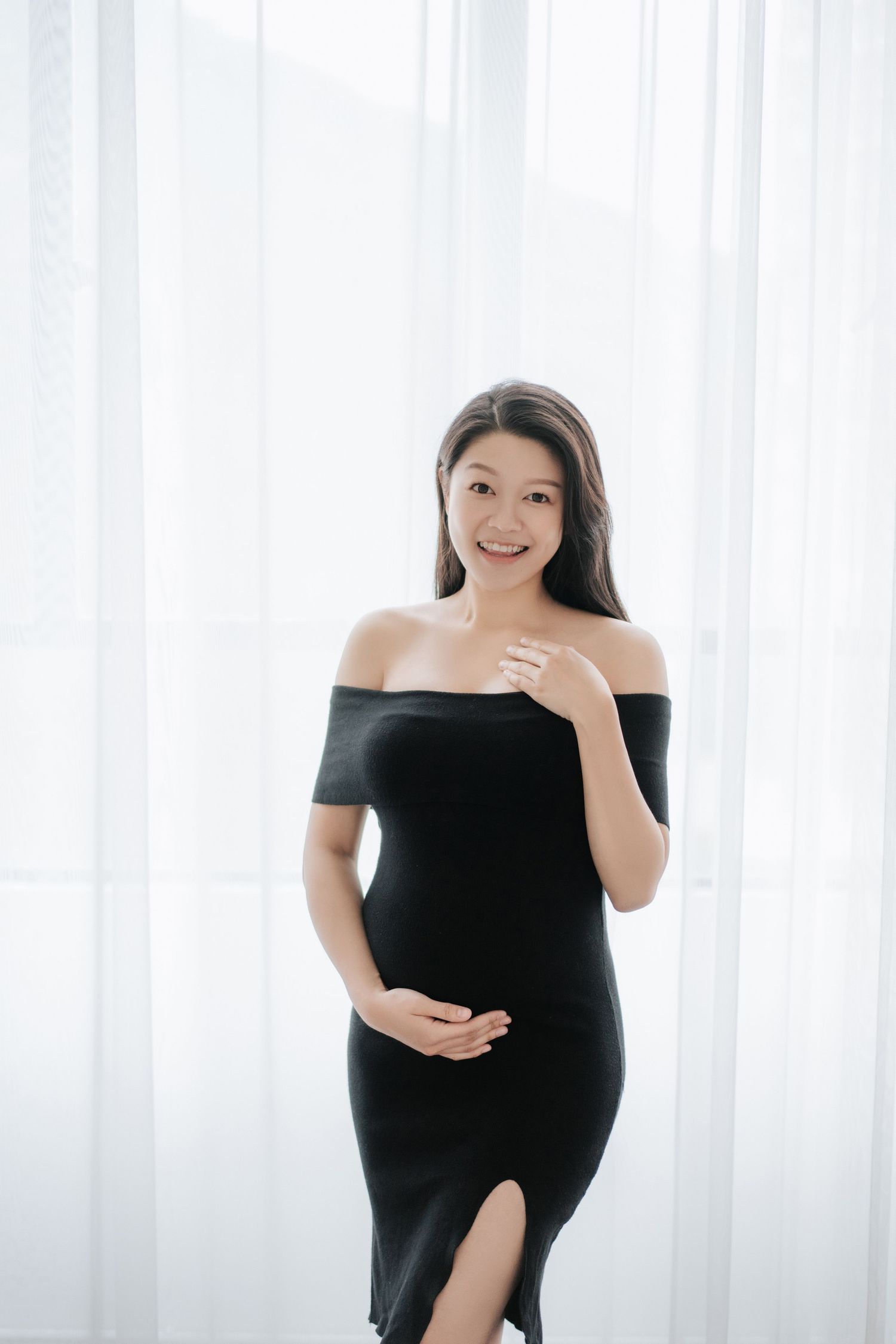 A maternity photo shoot featuring an off-shoulder black dress against a bright white curtained background.