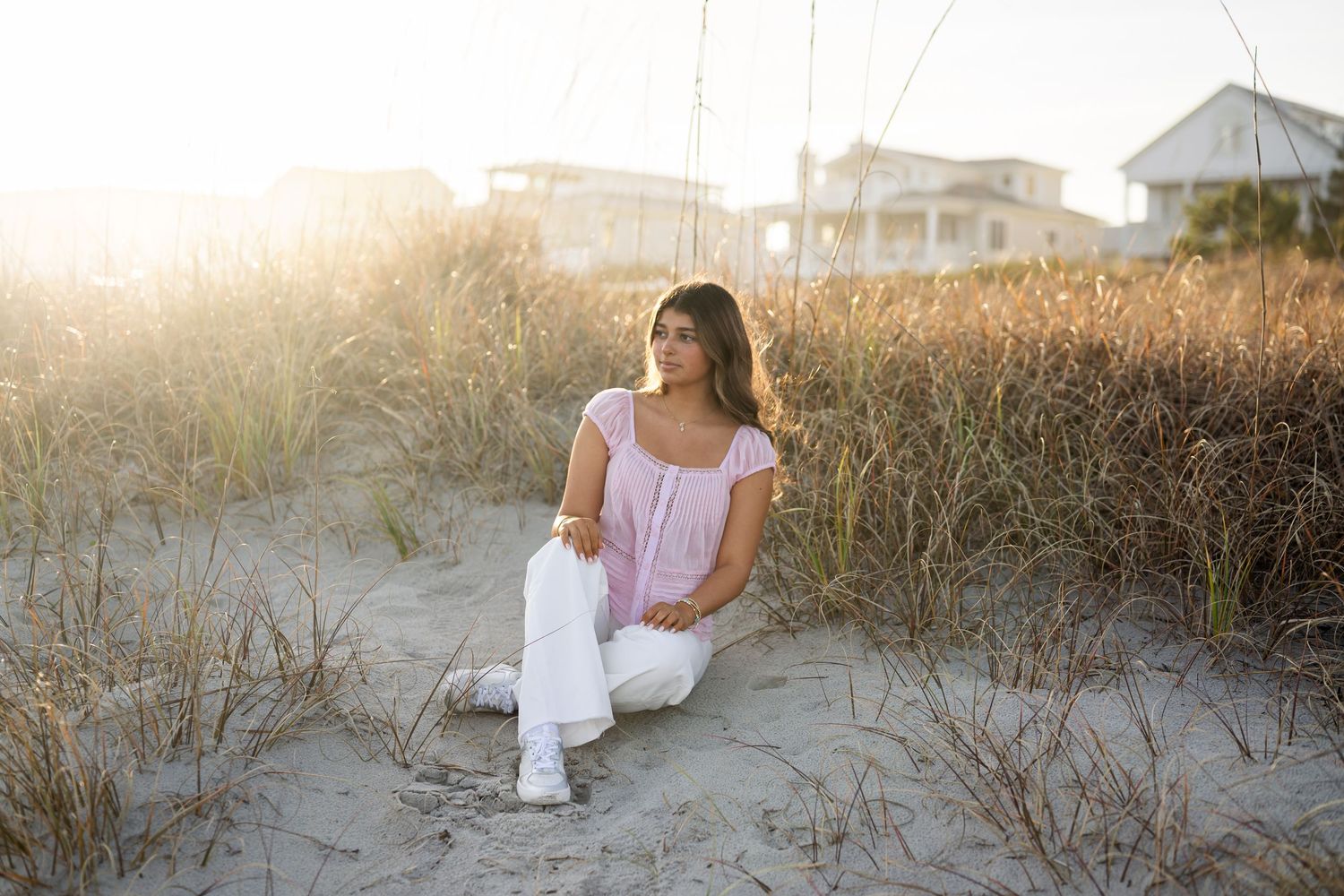 Figure in a white dress sits contemplatively by the ocean shore during sunset, creating a serene coastal scene.