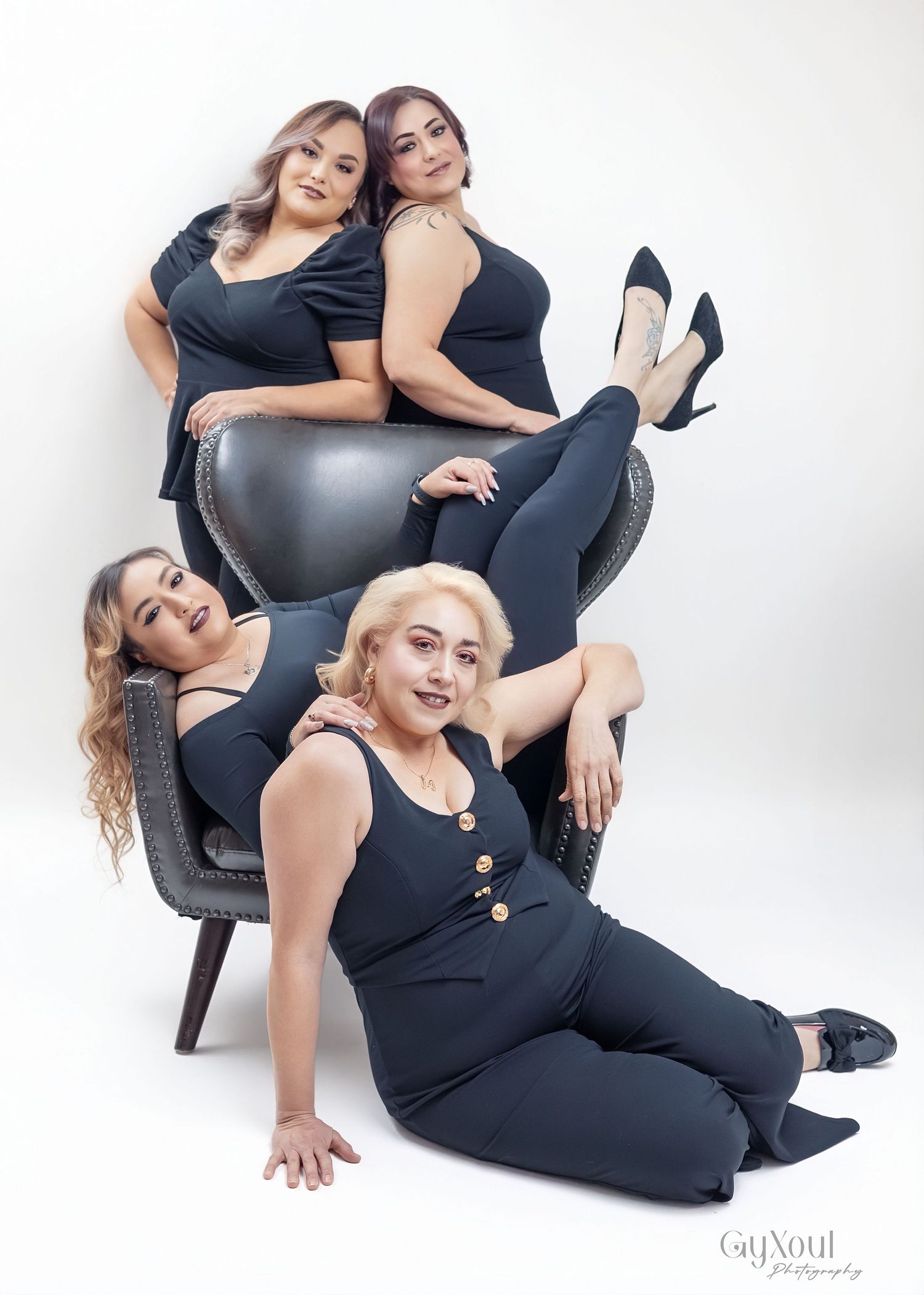 Two women in matching black outfits pose together on a gray chair for a fashion photoshoot series.