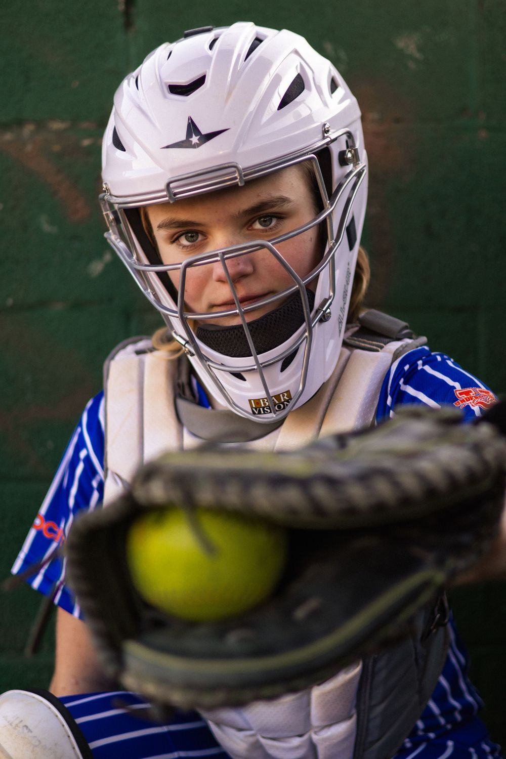 Softball catcher in white helmet and protective gear with yellow ball in frame.