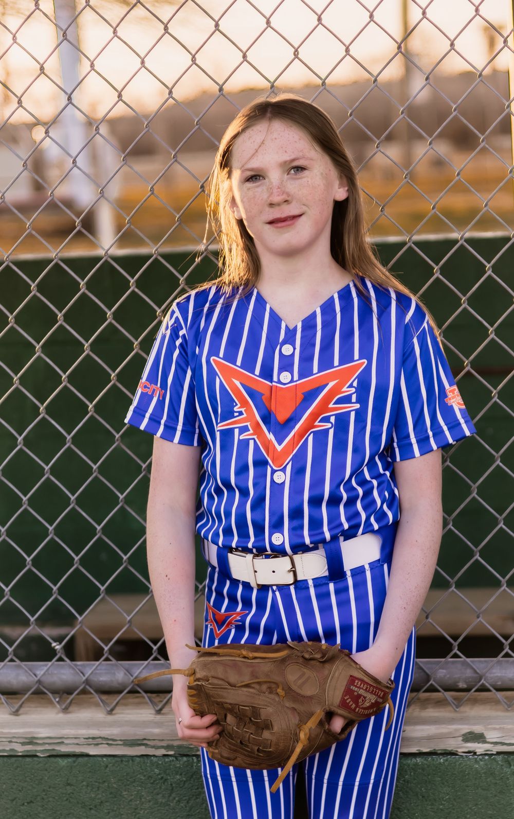 Player in blue pinstriped uniform posing with softball equipment by chain-link fence.