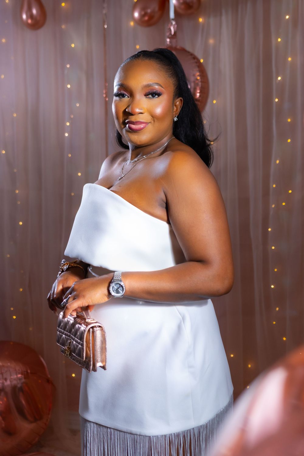 A figure in a white one-shoulder top and silver fringe skirt poses against a backdrop with fairy lights and balloons.