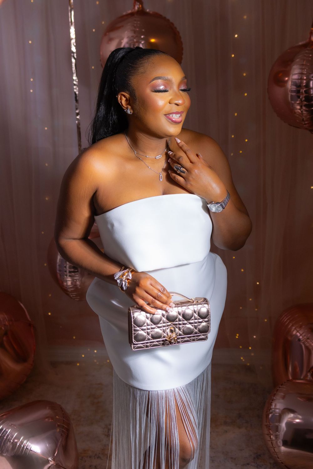 A woman in a white formal dress with a metallic clutch stands amidst rose gold balloon decorations.