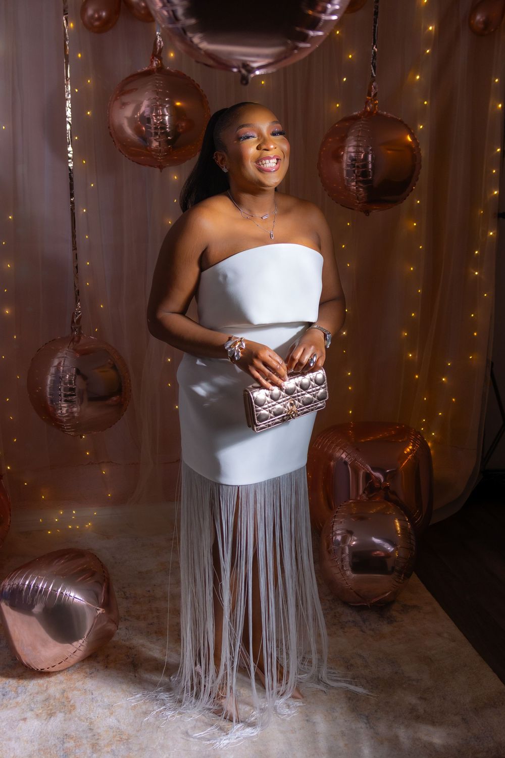 Elegant white gown with fringe detail and sparkly clutch against rose gold balloon and twinkle light backdrop.