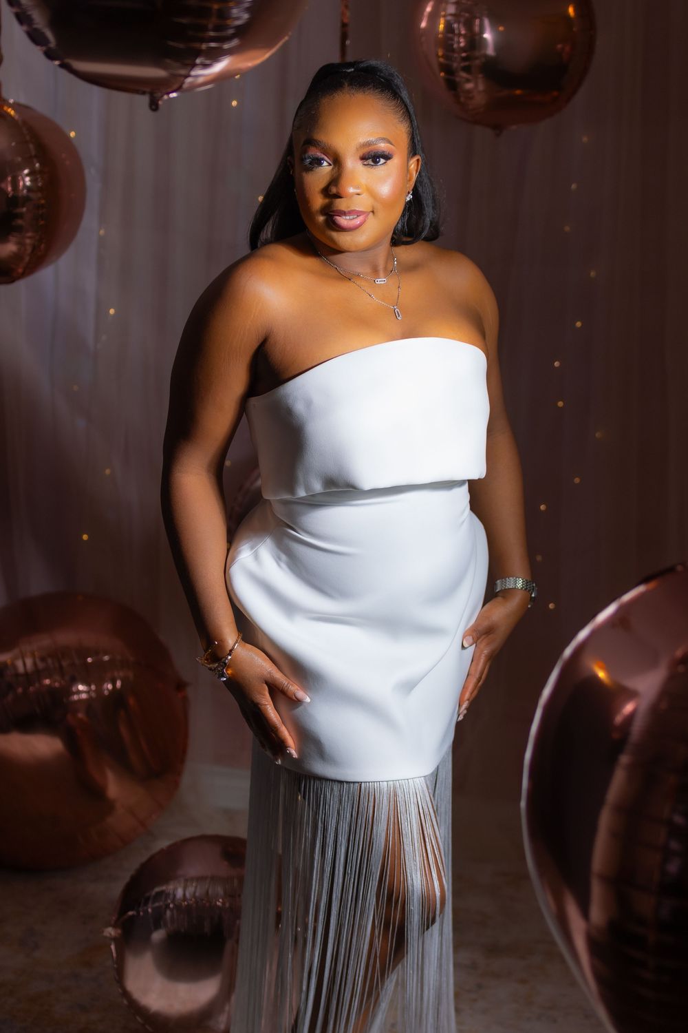 A model in a white asymmetrical top and silver fringe skirt poses elegantly with metallic balloon decorations.