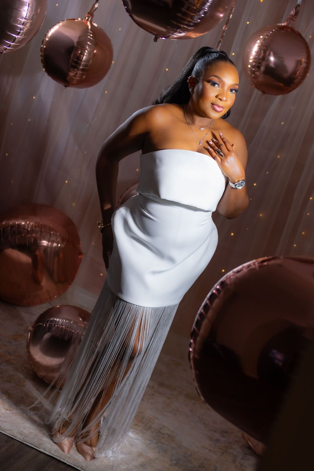 A person in a white strapless dress with a flowing gray skirt poses against a backdrop of brown balloon decorations.