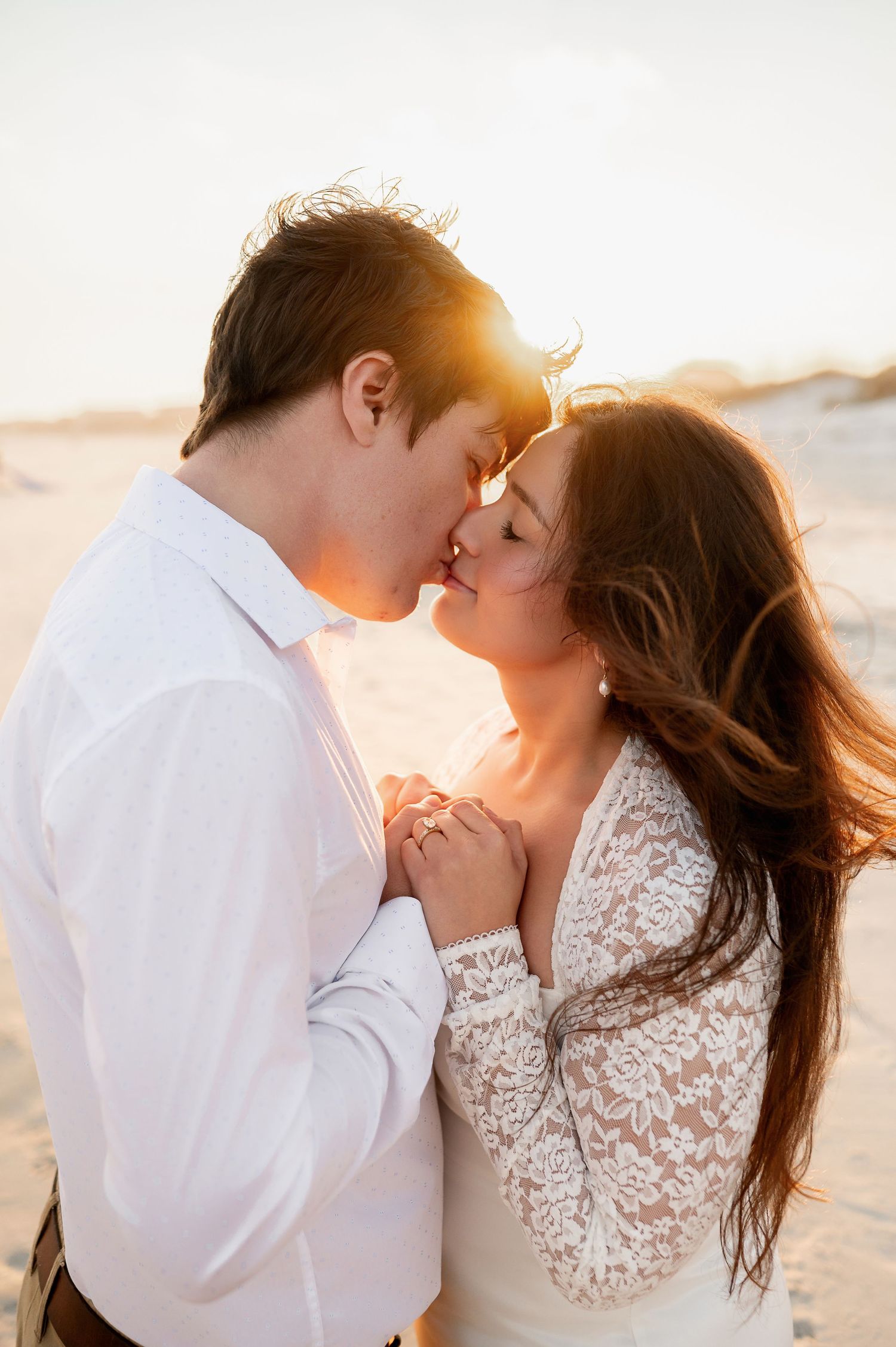 Romantic beach engagement photo of couple kissing in white attire during sunset.
