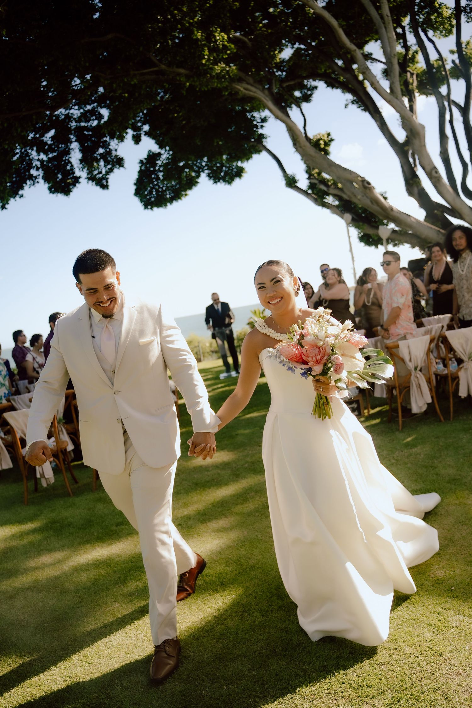 A romantic wedding couple embraces outdoors with tropical florals and greenery in an elegant Hawaiian ceremony setting.