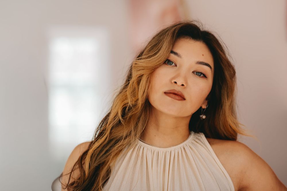 A person with flowing caramel-colored hair poses in a cream halter top against a bright, softly lit background.