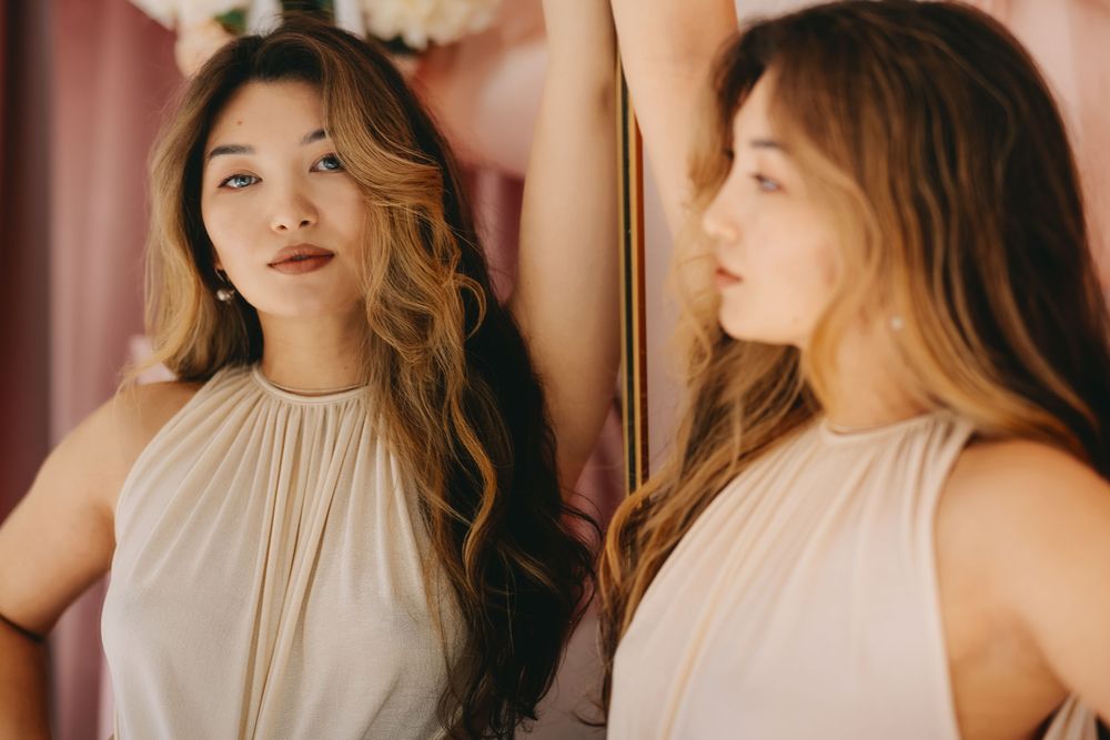 Woman in beige halter dress poses next to mirror reflection in warm lighting.