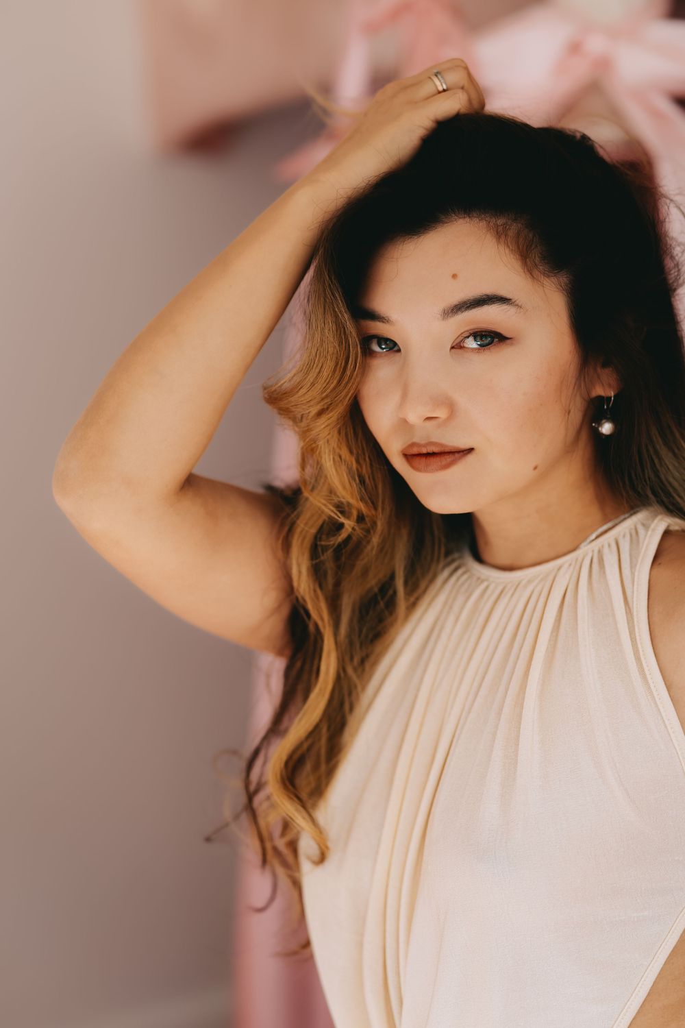 A portrait of a woman in a cream-colored sleeveless top with long wavy ombre hair posing against a pink background.