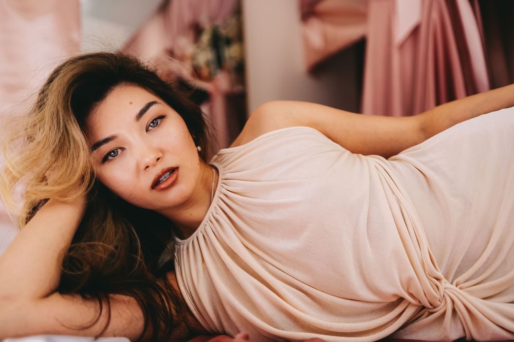 A person in a blush-colored dress poses elegantly on a pink couch in warm indoor lighting.