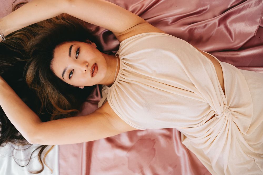 A person with long dark hair lies on pink satin sheets wearing a cream colored dress in a romantic boudoir setting.