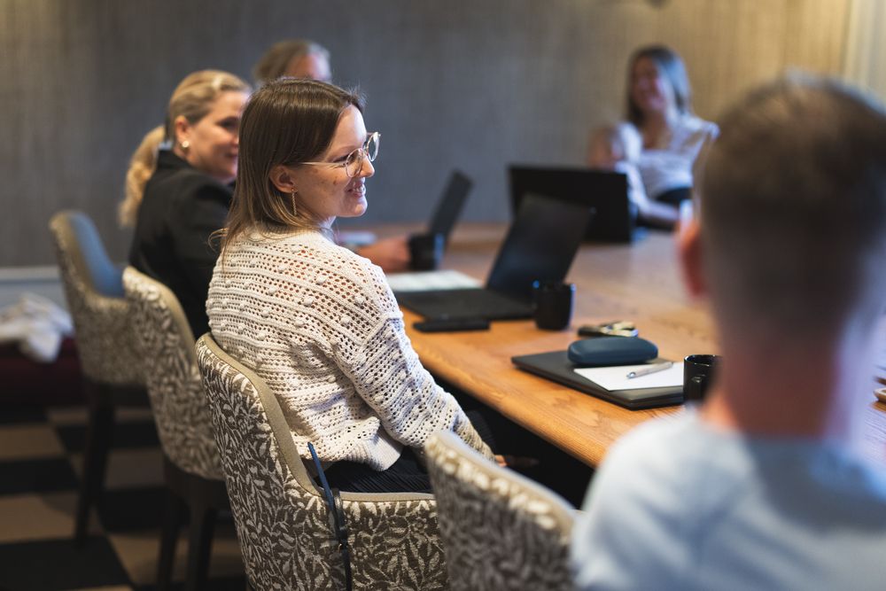 People sit around a conference table with laptops during a business meeting in a modern office setting.