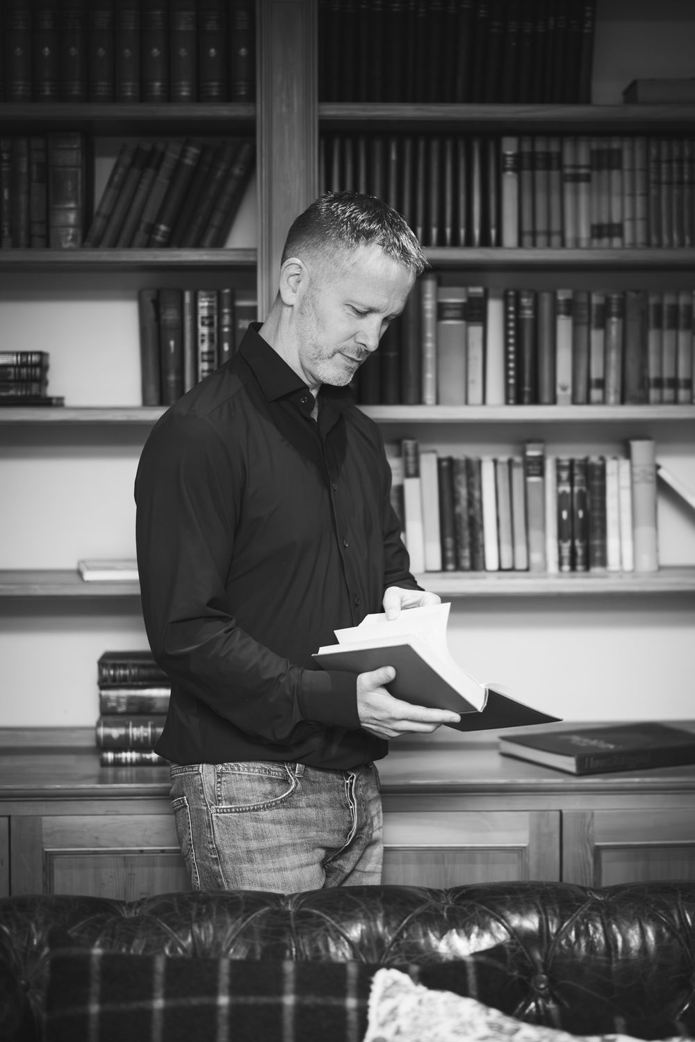 Person in black shirt reading on leather couch with bookshelf in background.