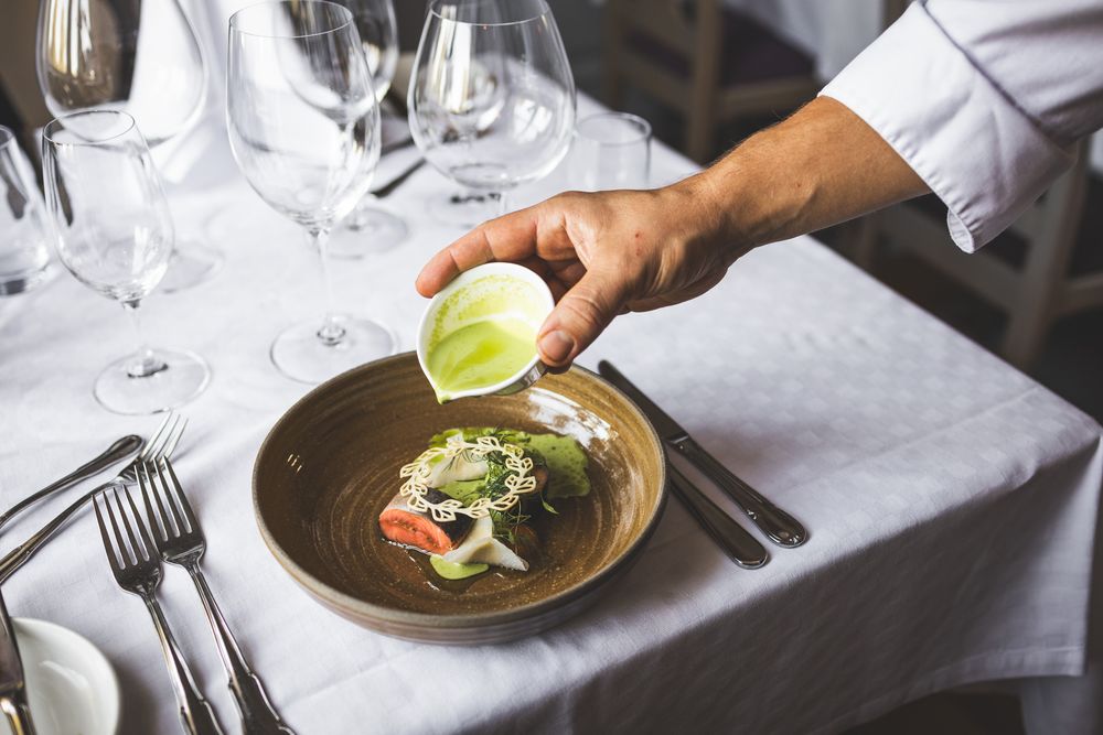 Chef squeezing lime juice over a plated salad at an elegant restaurant table set with wine glasses.