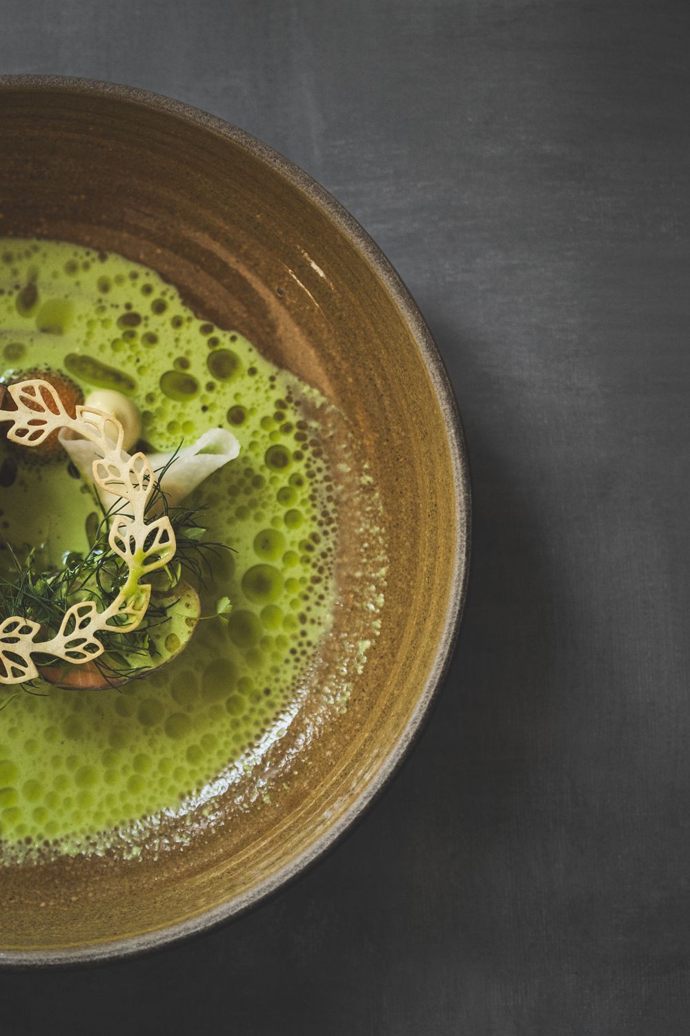 A vibrant green pea soup with delicate leaf-shaped garnishes served in a rustic brown ceramic bowl against dark background.