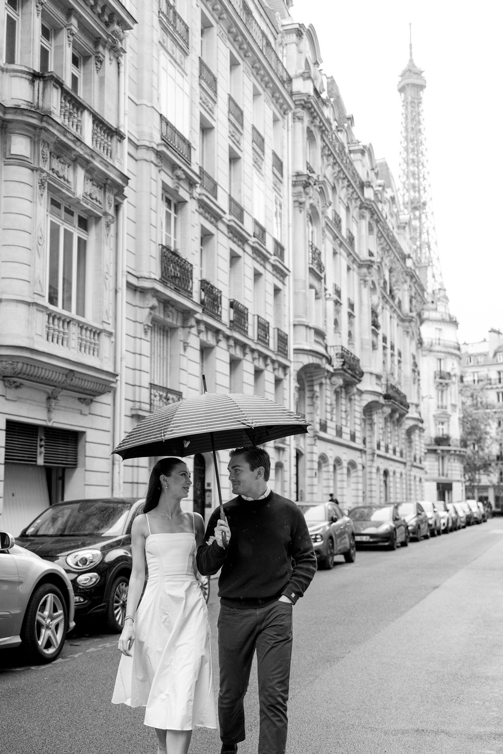Couple sharing umbrella on romantic Paris street with Eiffel Tower visible in background among classic architecture.