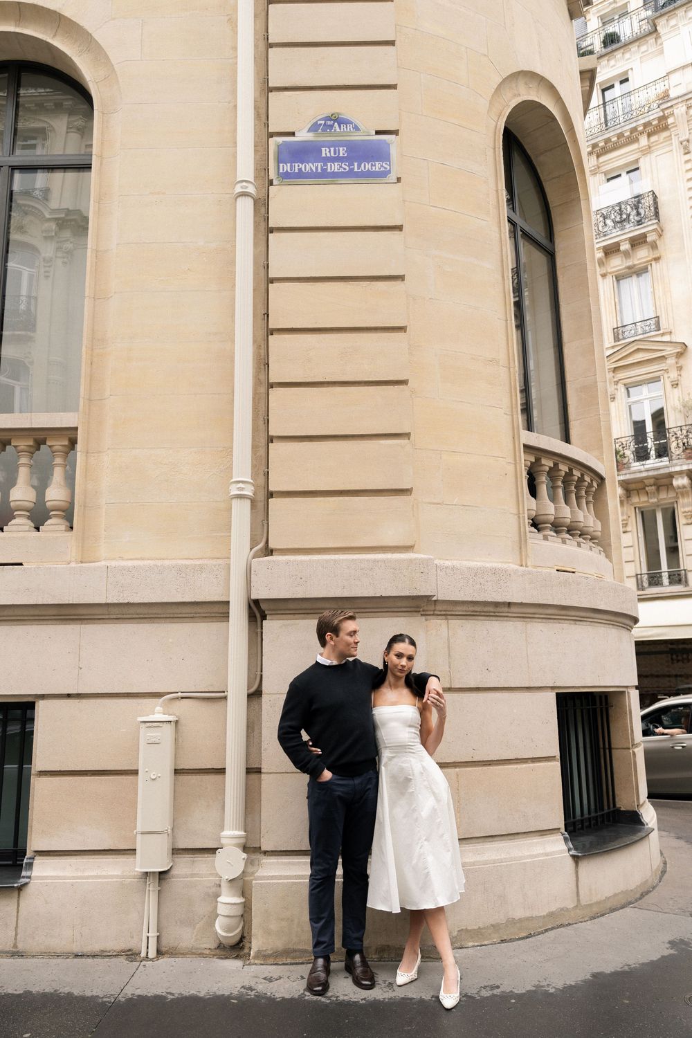 Couple in formal attire standing on a Parisian street corner with classic architecture and a blue street sign.