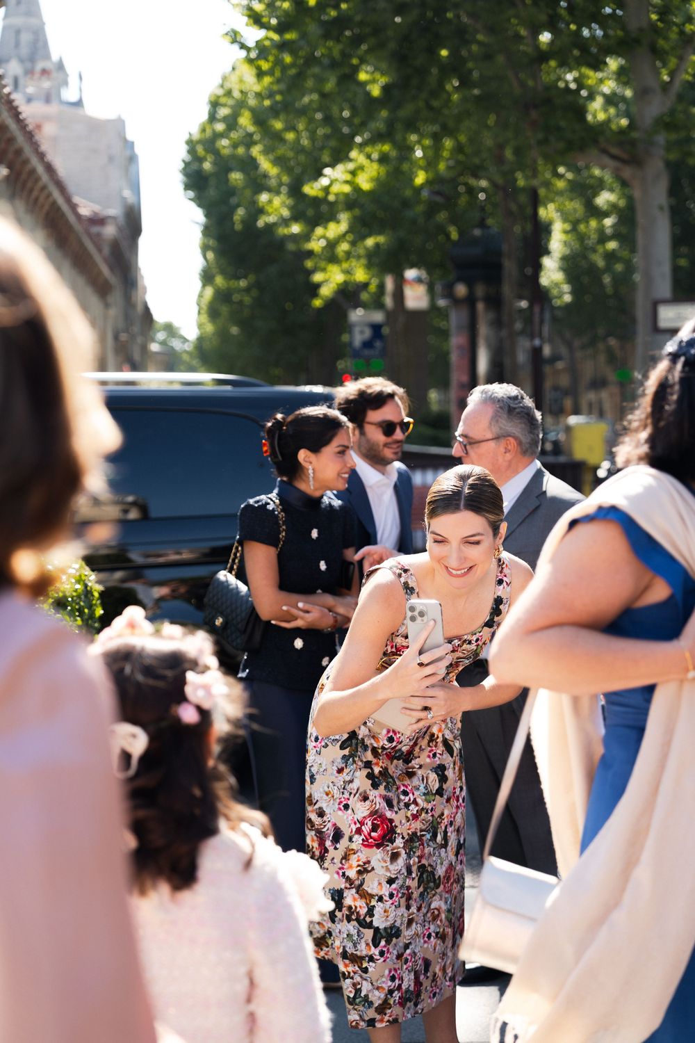 A group of wedding guests gather outdoors on a sunny day with trees in the background.