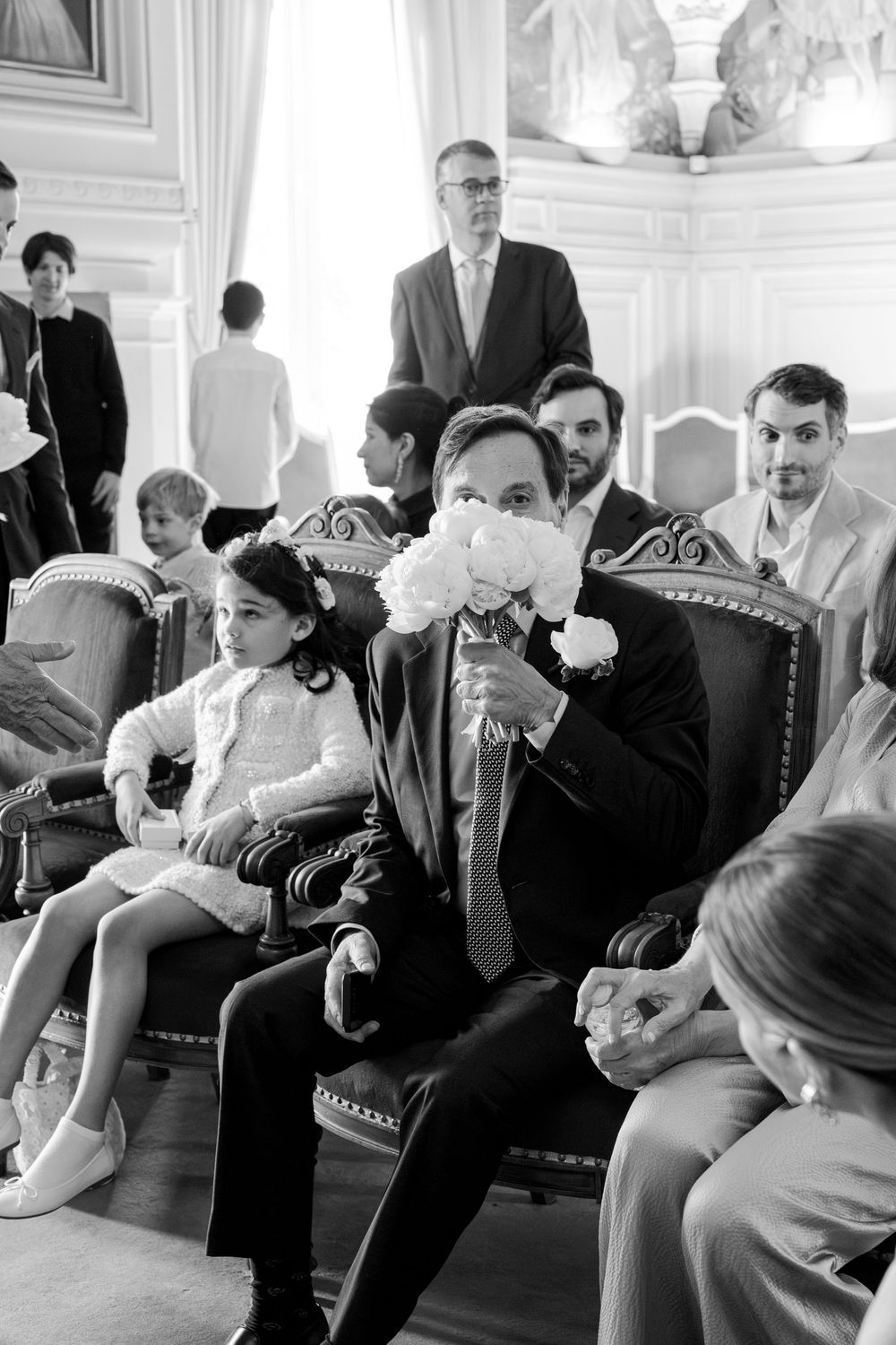 Black and white photo of guests seated on ornate chairs in an elegant room with someone smelling a white flower bouquet.