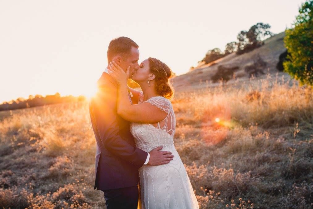 A couple embraces during golden hour on a hillside as the sun sets behind them creating a warm silhouette.