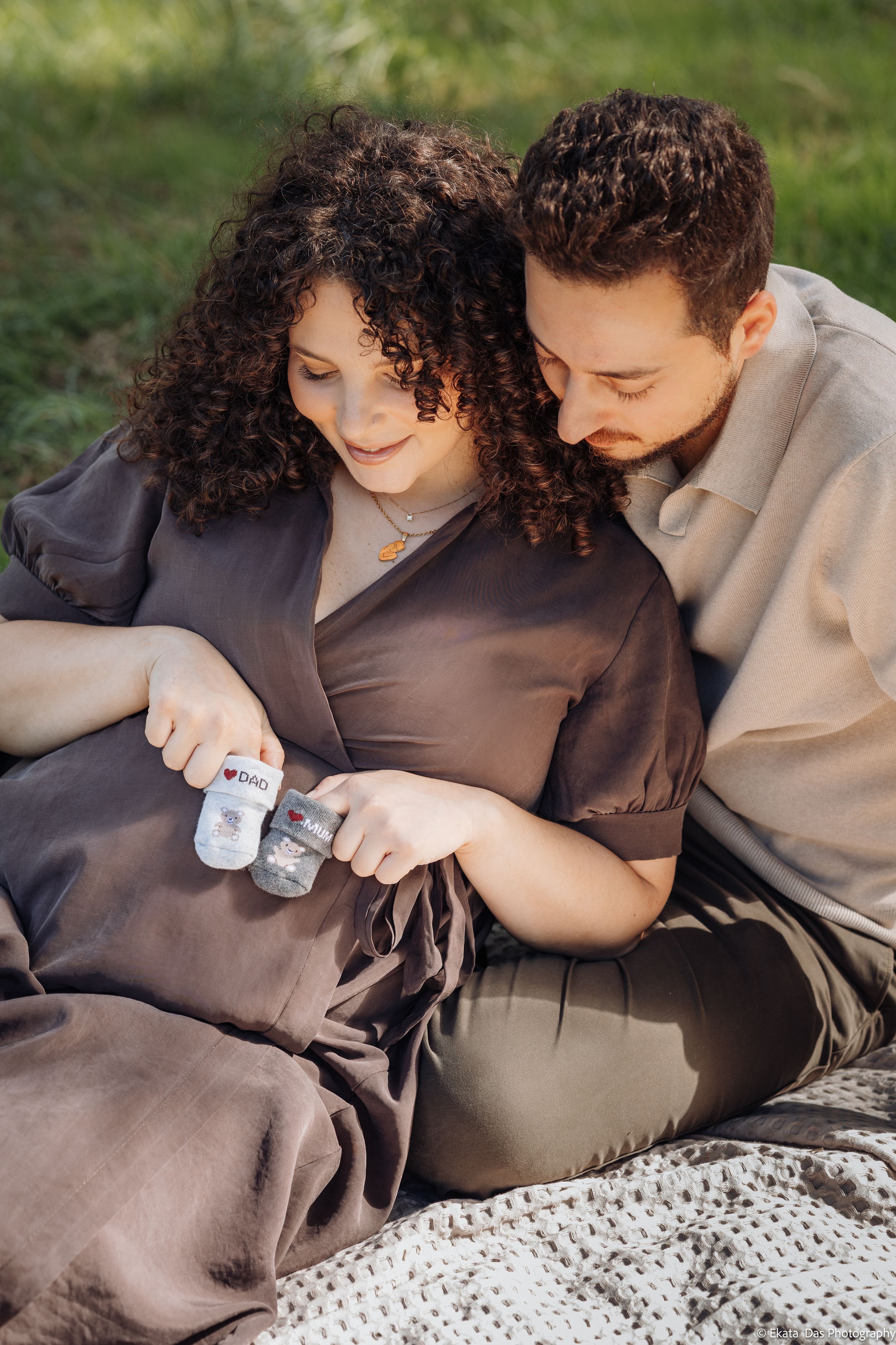 Expecting couple sitting on a blanket in the grass as the pregnant woman holds tiny baby booties over her belly while both smile down.