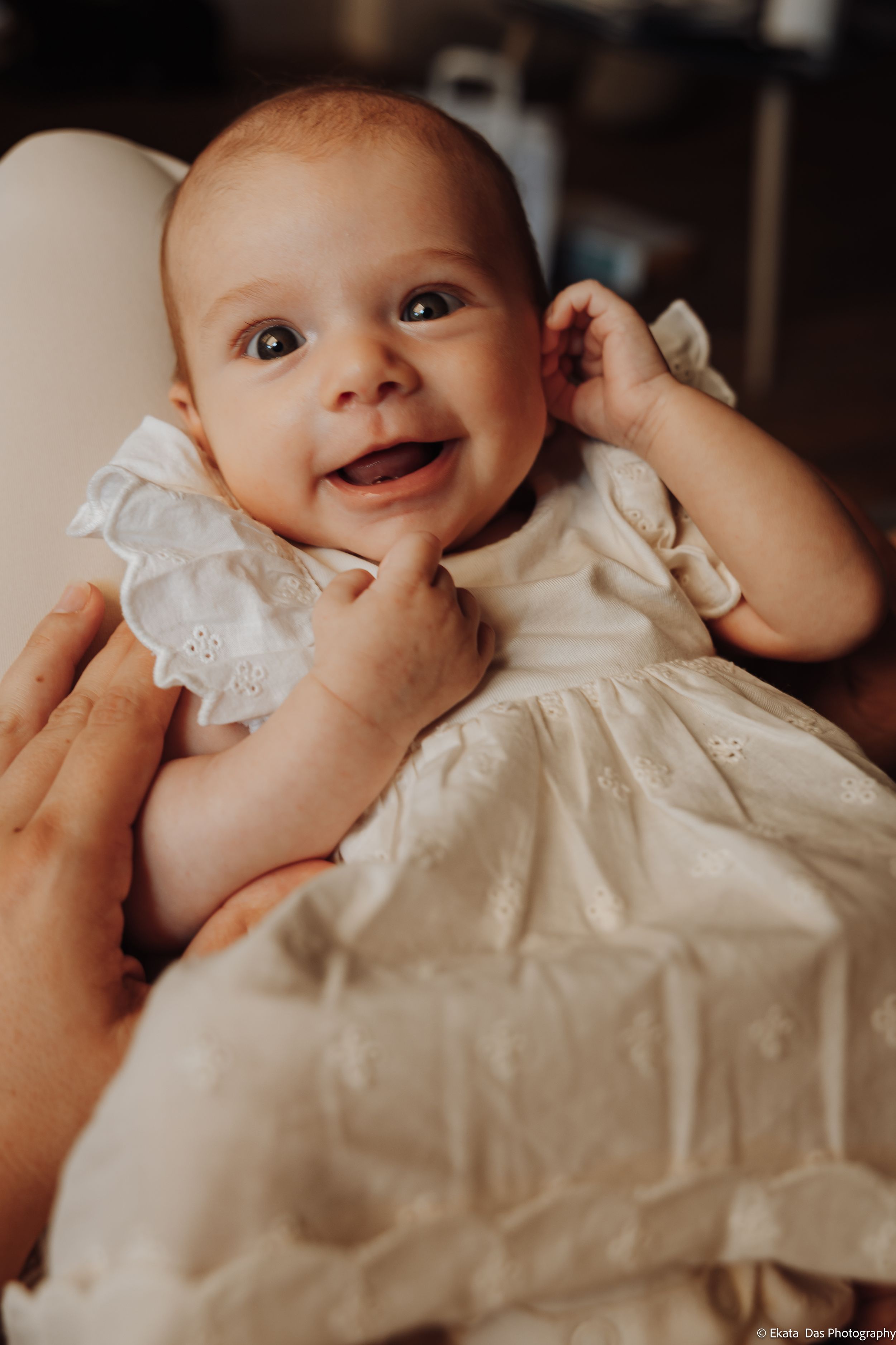 Smiling baby in a white eyelet lace dress with ruffled sleeves being held in an adult's arms, with hand raised toward the chin.