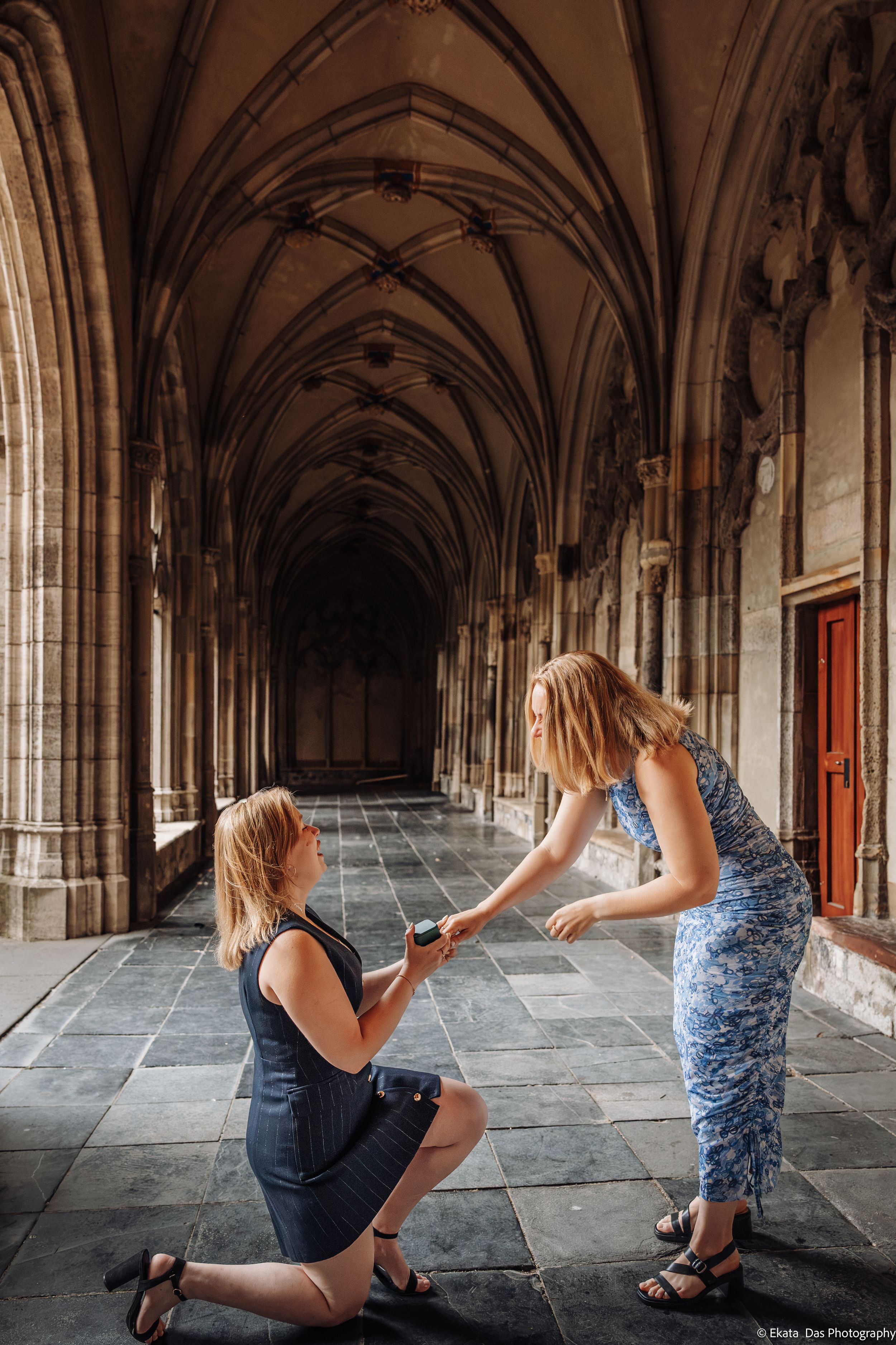 Woman in a dark dress kneels with a ring box proposing to her partner in a blue floral dress in a historic Gothic cloister.